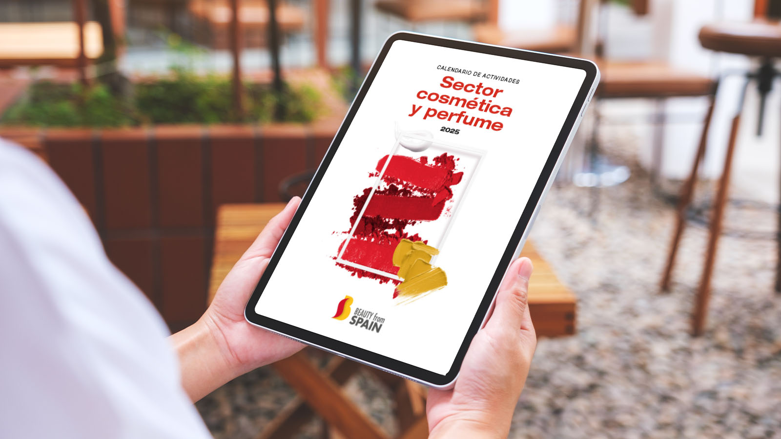Una persona sostiene una tablet donde puede ver la portada del calendario de actividades de ICEX para el sector de perfumería y cosmética