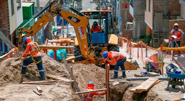 Varios obreros y una excavadora trabajan sobre un zanja realizada en la vía pública.