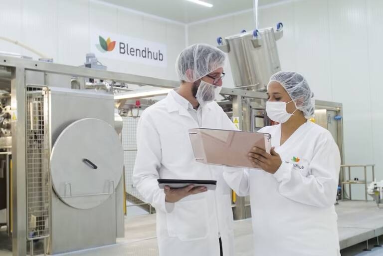 Dos personas uniformados de blanco, con mascarilla y gorro charlan acerca de un documento en las instalaciones de una fábrica o labotorio.