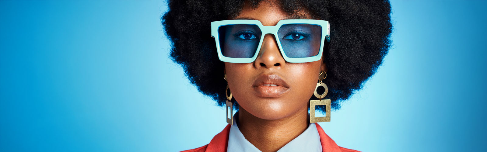 Una mujer de raza negra con pelo a lo afro, unas gafas azul verdoso y unos pendientes dorados.