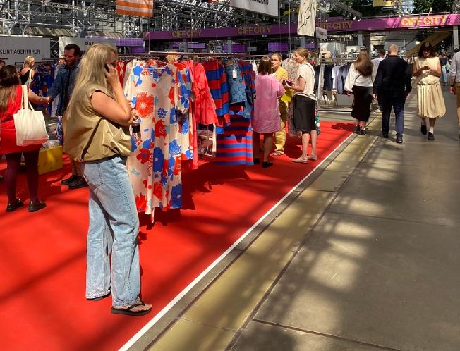 Una mujer rubia habla por teléfono al lado de una exposición de vestidos de mujer, en un stand con moqueta roja lleno de gente, dentro del Pabellón España de CIFF