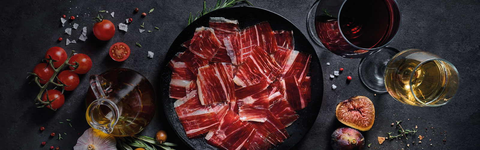 Bodegón con tomates, aceite, jamón ibérico, dos copas de vino tinto y blanco y más alimentos sobre un fondo oscuro