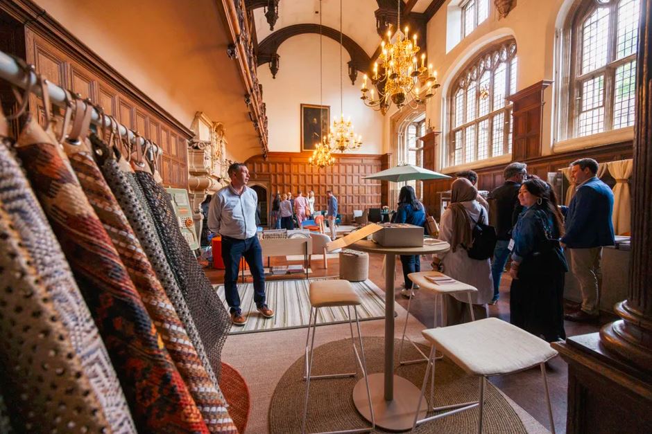 Una sala de exposiciones en el museo de la Orden de San Juan, con un store de alfombras, una mesa, varias sillas y varios visitantes viendo otros muebles de diseño de la exposición.