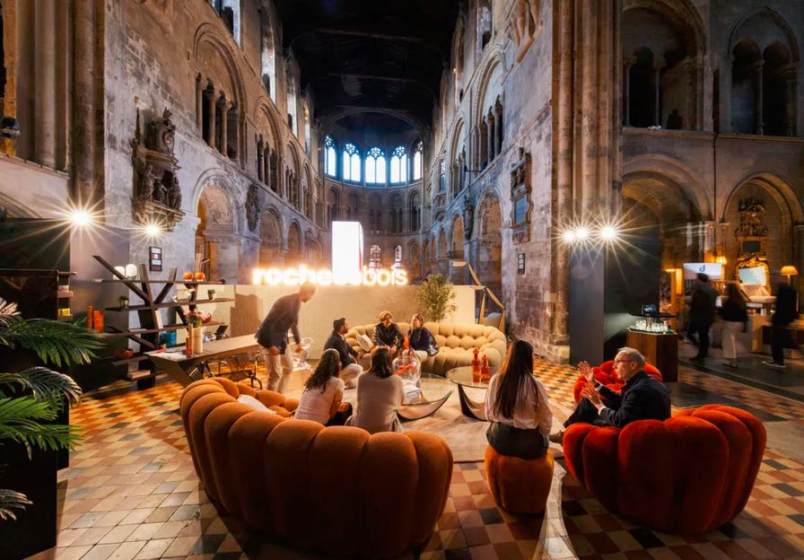 Varias personas se sientan en sofás y asientos de diseño. Detrás se ve un estrado con un cartel luminoso, y, al fondo, la arquitectura del museo de la Orden de San Juan.