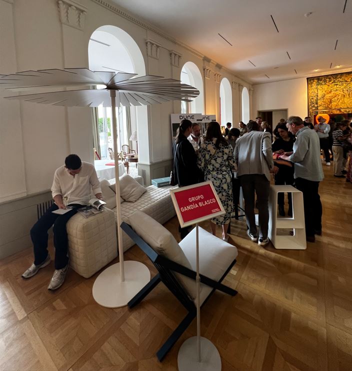 Una persona mira un catálogo sentado en un sofá de diseño, y detrás, varios visitantes se congregan en frente de una exposición