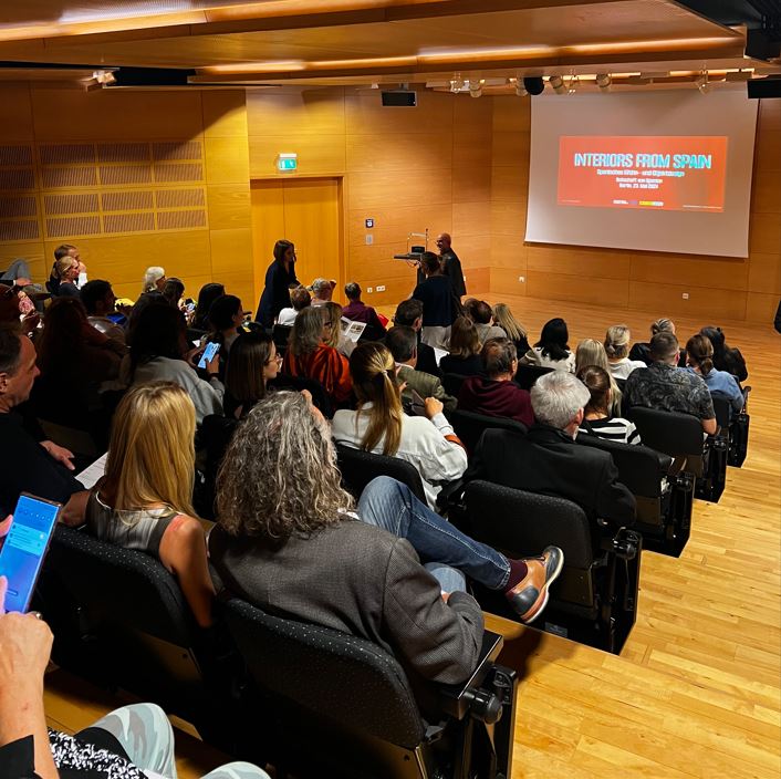 Varias personas ocupan 5 filas de butacas negras y atienden a la conferencia de un ponente, que da la ponencia con una presentación en un proyector.