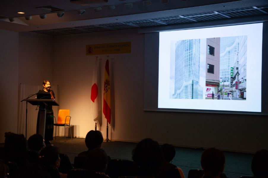 Una mujer asiática realiza una presentación sobre un escenario, detrás tiene las banderas de España y Japón, y a su derecha, un proyector donde se ve la fachada de un edificio en la calle