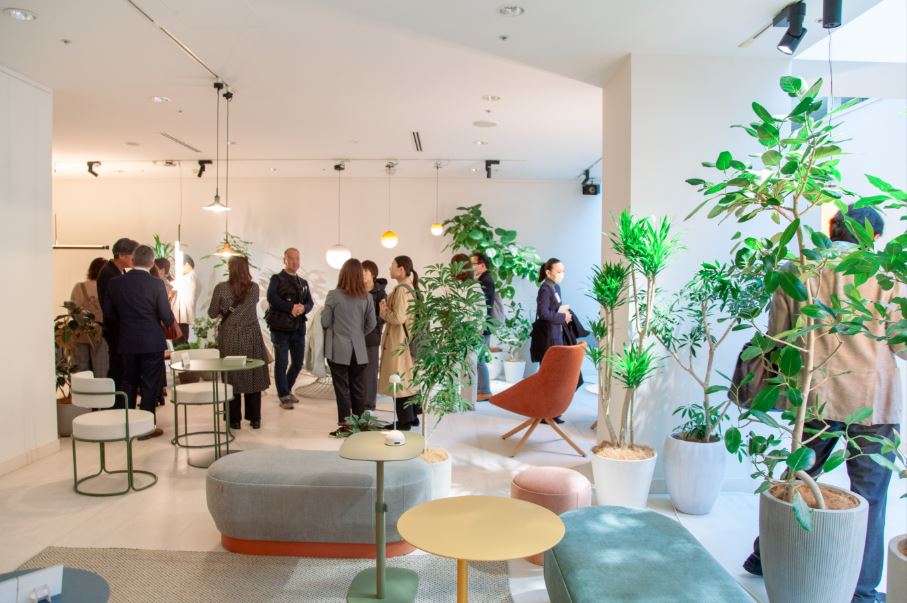 Una sala de exposición con plantas, muebles y textil de hogar, al fondo se ven varios visitantes hablando y observando lámparas de iluminación