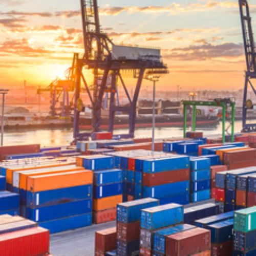 Varios contenedores marítimos, detrás de ellos una grúa, y detrás, el atardecer.