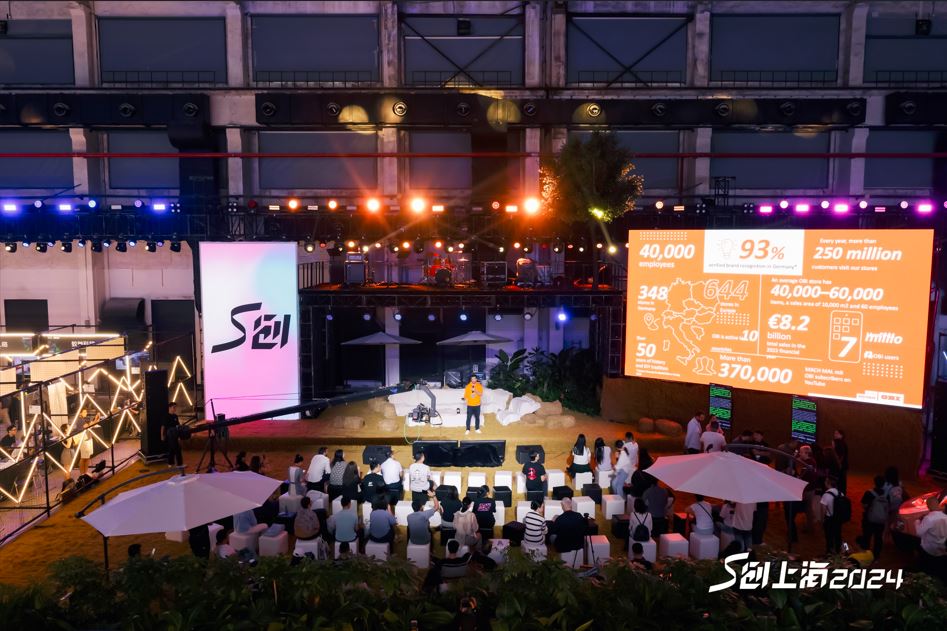 Un conferenciante da una ponencia en un escenario ambientado con arena de playa y sombrillas blancas, frente a un público de docenas de personas.