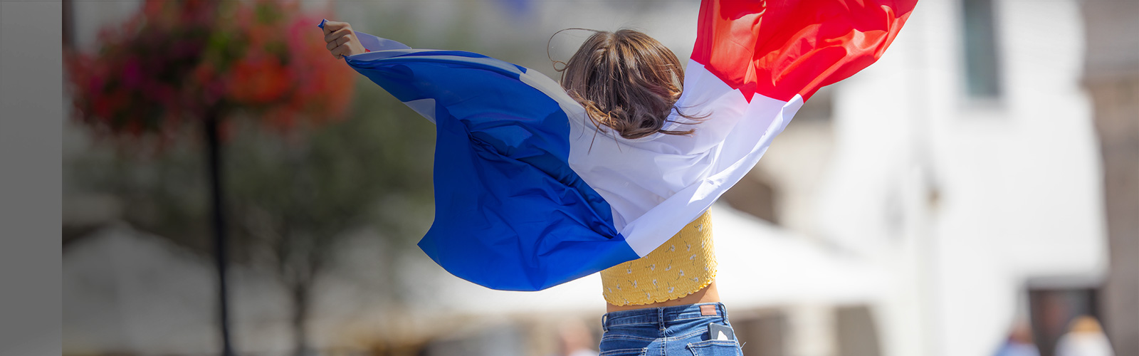 Una niña rubia de espaldas, con camiseta amarilla y vaqueros, lleva sobre los hombros una bandera de Francia.