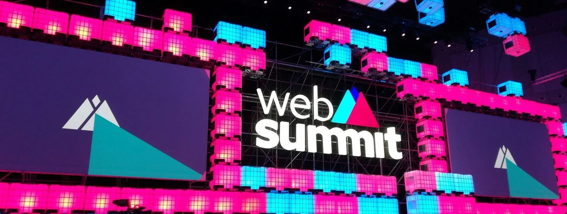 Un escenario de Web Summit, con paneles de luz rosas y azules, y varias pantallas en el medio