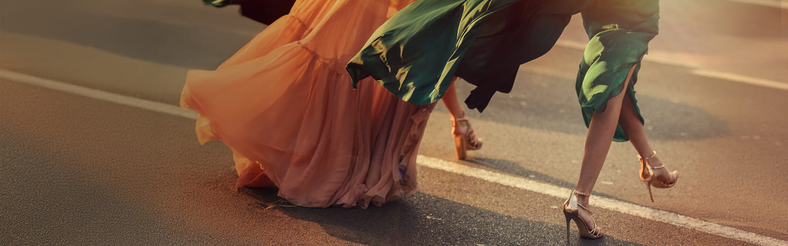 Dos mujeres de cintura para abajo caminan, una de ellas lleva un vestido verde, y la otra, a su izquierda, un vestido naranja