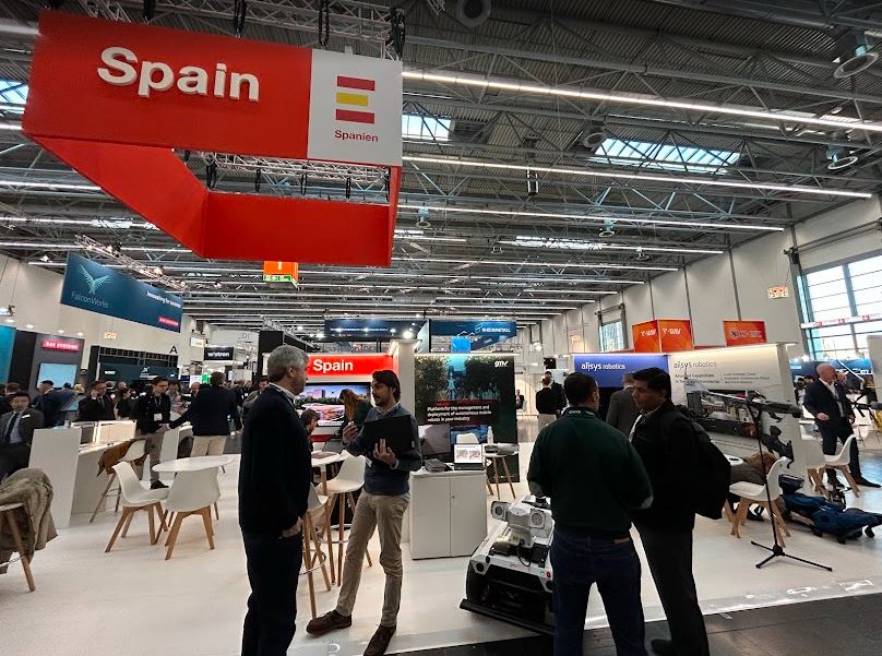 Dos grupos de dos personas conversan entre sí delante de la zona común de reuniones del Pabellón España en Xponential Europe.