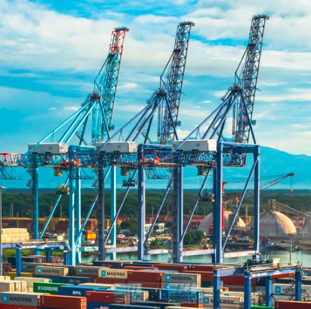 Tres grúas elevan varios contenedores marítimos, rodeadas de más contenedores marítimos en un puerto de mercancías.