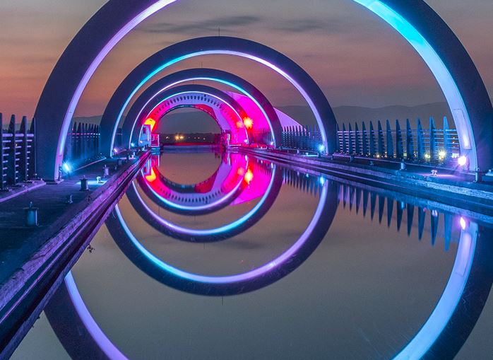 Un puente con aros grandes con iluminación moderna de colores. En el medio del puente hay un estanque de agua transparente.
