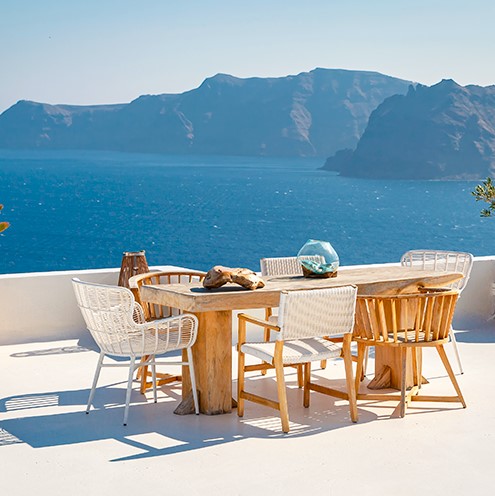 Una mesa de madera de terraza con cuatro sillas de madera alrededor, en una terraza blanca con vistas a la playa.