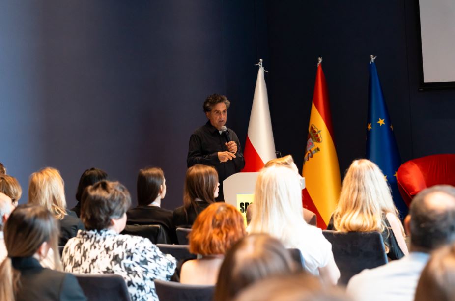Un arquitecto español imparte una ponencia ante decenas de prescriptores polacos. Al lado del ponente hay una bandera de Polonia, otra de España y otra de la UE.