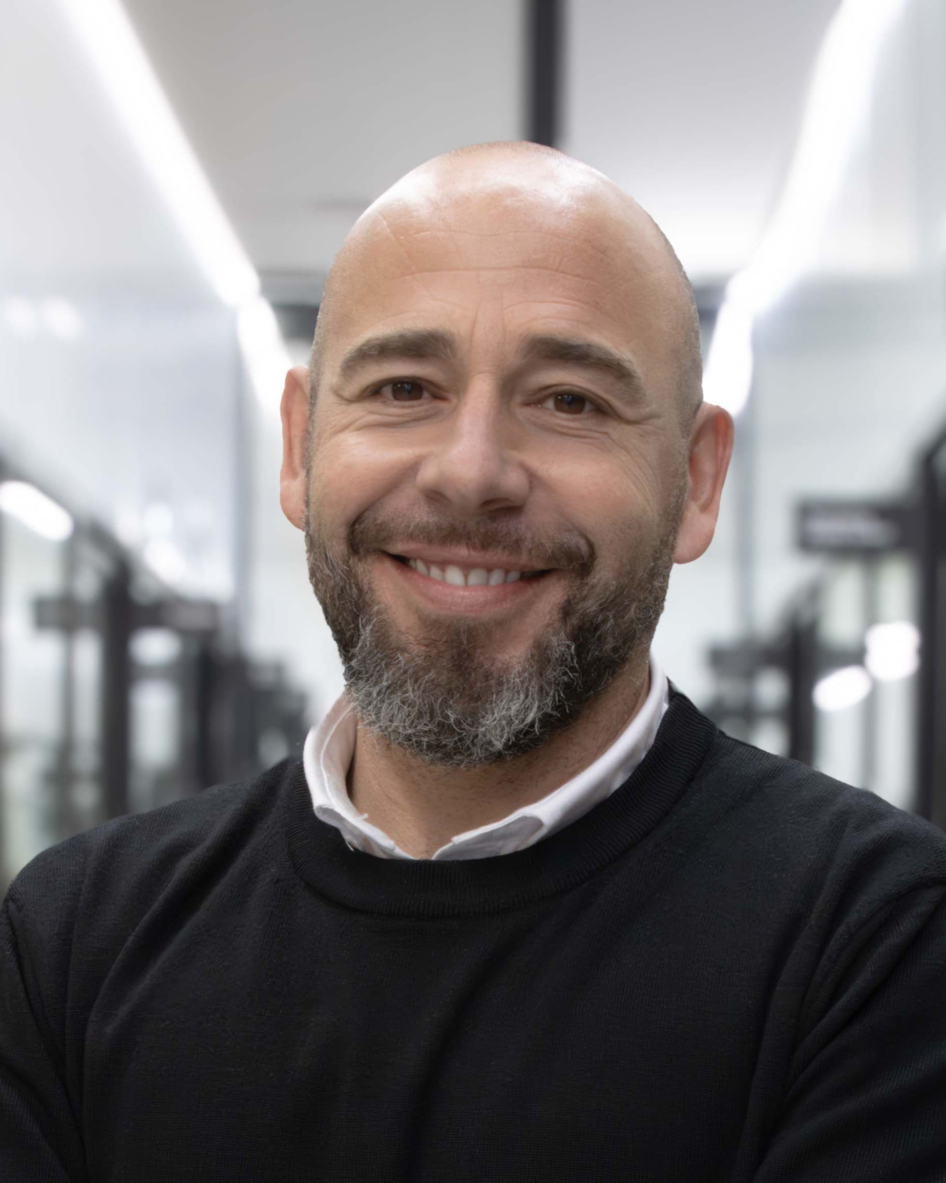 Hombre sonriendo a la cámara, con cabeza afeitada y barba entrecana, vestido con jersey negro y camisa blanca, en una oficina moderna desenfocada.