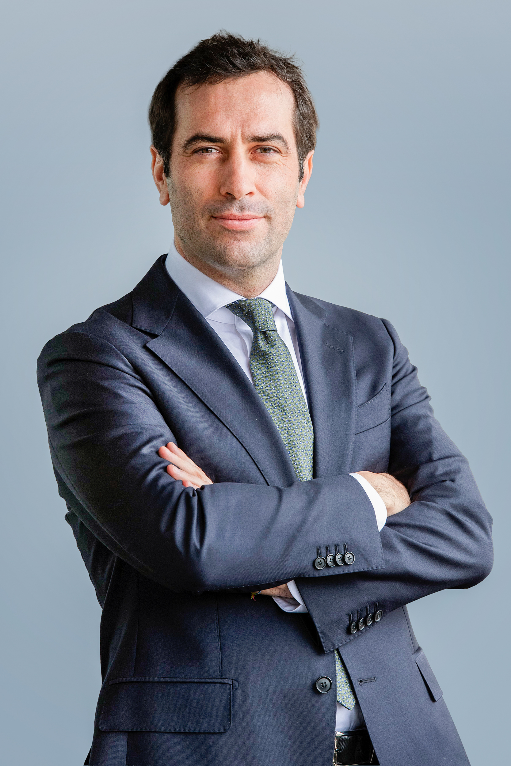 Hombre con traje oscuro y corbata clara, posando con los brazos cruzados frente a un fondo gris en un retrato corporativo