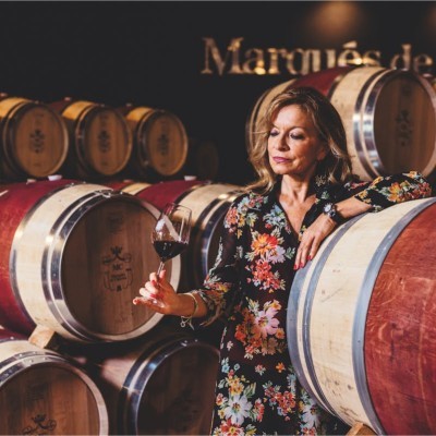 Mujer en una bodega rodeada de barricas de madera, sosteniendo una copa de vino tinto. La imagen refleja la excelencia, la tradición y la proyección internacional del sector vitivinícola español.