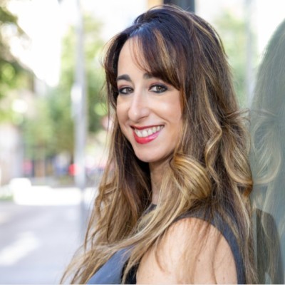 Retrato en primer plano de una mujer con cabello largo y ondulado, vestida con prenda oscura. Fondo exterior desenfocado con vegetación y luz natural. La iluminación destaca los rasgos y la sonrisa.