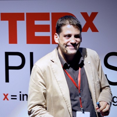 Persona sobre un escenario durante una charla TEDx, con micrófono de diadema y acreditación colgada al cuello. Al fondo destaca el logo rojo “TEDx” sobre una pantalla blanca iluminada