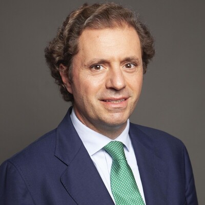 Retrato en color de un hombre encuadrada desde el pecho hacia arriba, con el cuerpo ligeramente girado y el rostro orientado hacia la cámara. Lleva traje azul, camisa blanca y corbata verde con pequeños puntos. El fondo es neutro gris, con iluminación suave que define con claridad los rasgos y el peinado ondulado.