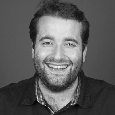 Retrato en blanco y negro de una persona con barba y cabello corto, sonriendo con dientes visibles. Viste camisa oscura sobre otra de cuadros. Fondo liso gris, iluminación suave y encuadre cercano.