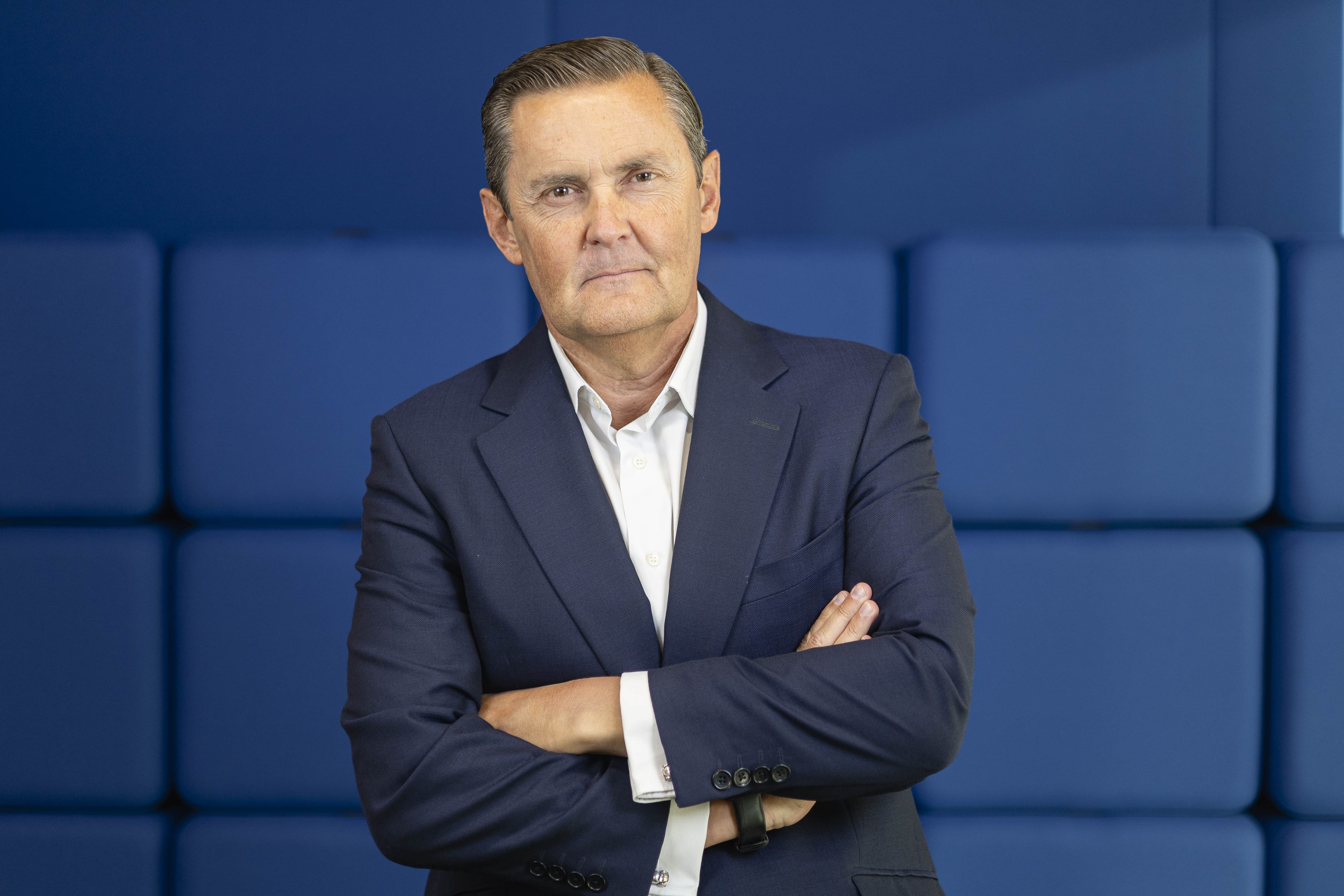 Hombre con traje azul marino y camisa blanca, de pie con los brazos cruzados frente a un fondo azul acolchado, mirando a cámara con expresión seria y profesional.