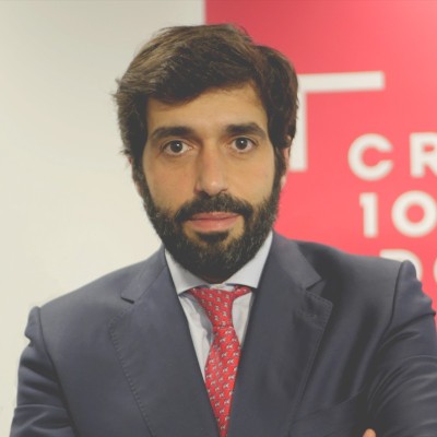 Hombre con barba y cabello oscuro, vestido con traje azul marino, camisa blanca y corbata roja, mirando a la cámara en un entorno interior.
