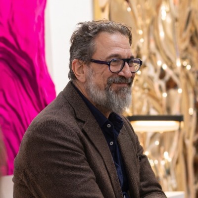 Hombre con barba y gafas, vestido con chaqueta marrón, sentado en un espacio interior con iluminación cálida y fondo artístico.