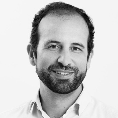 Retrato en blanco y negro de un hombre en primer plano, centrado desde los hombros hacia arriba. Presenta cabello corto y barba recortada. Lleva camisa clara con el cuello abierto. El fondo es liso y claro, con iluminación uniforme que resalta los rasgos faciales y la textura del cabello.