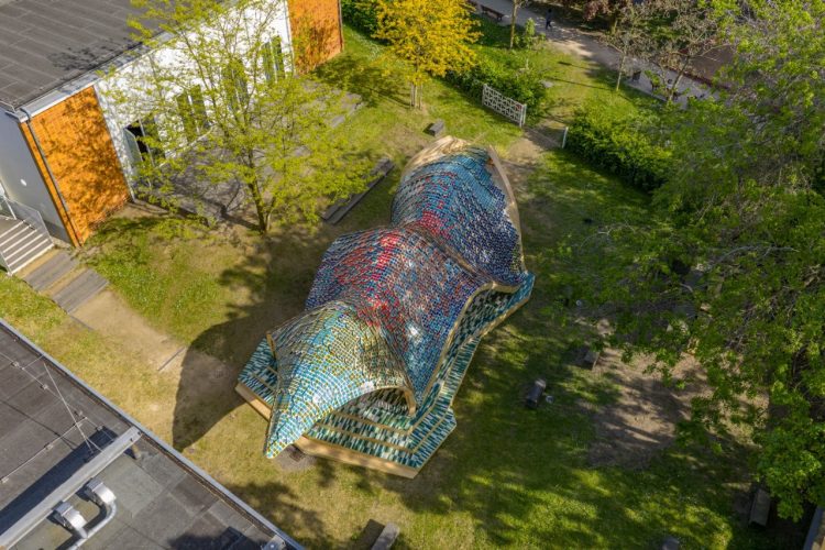 Foto panorámica del escenario donde se va a celebrar la exposición, se ve su arquitectura moderna con tejado con mosaicos azules y rojos., y a su alrededor un jardín con árboles y unos edificios.