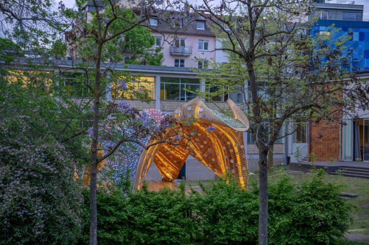 Imagen del escenario escondido detrás de dos árboles y unos arbustos del jardín, se puede ver la fachada de su arquitectura con paredes amarillas y tejado azul y rojo.