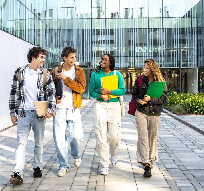 Cuatro alumnos caminan en los exteriores de un campus, con dos facultades de fachadas acristaladas alrededor.