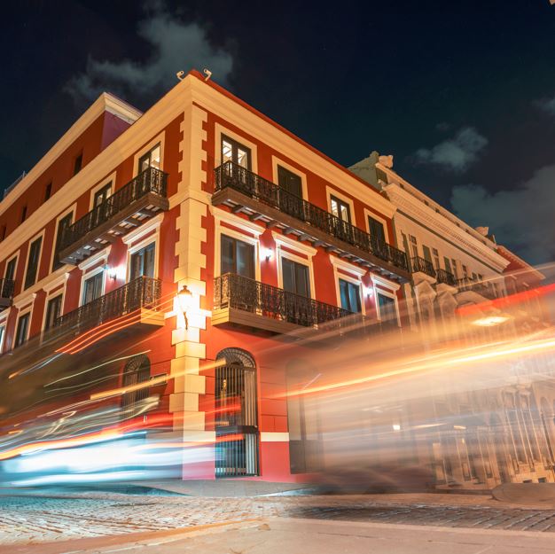 Estelas distorsionadas que reproducen el movimiento de coches y personas, y detrás, un edificio de fachada clásica roja con ventanas y balcones.