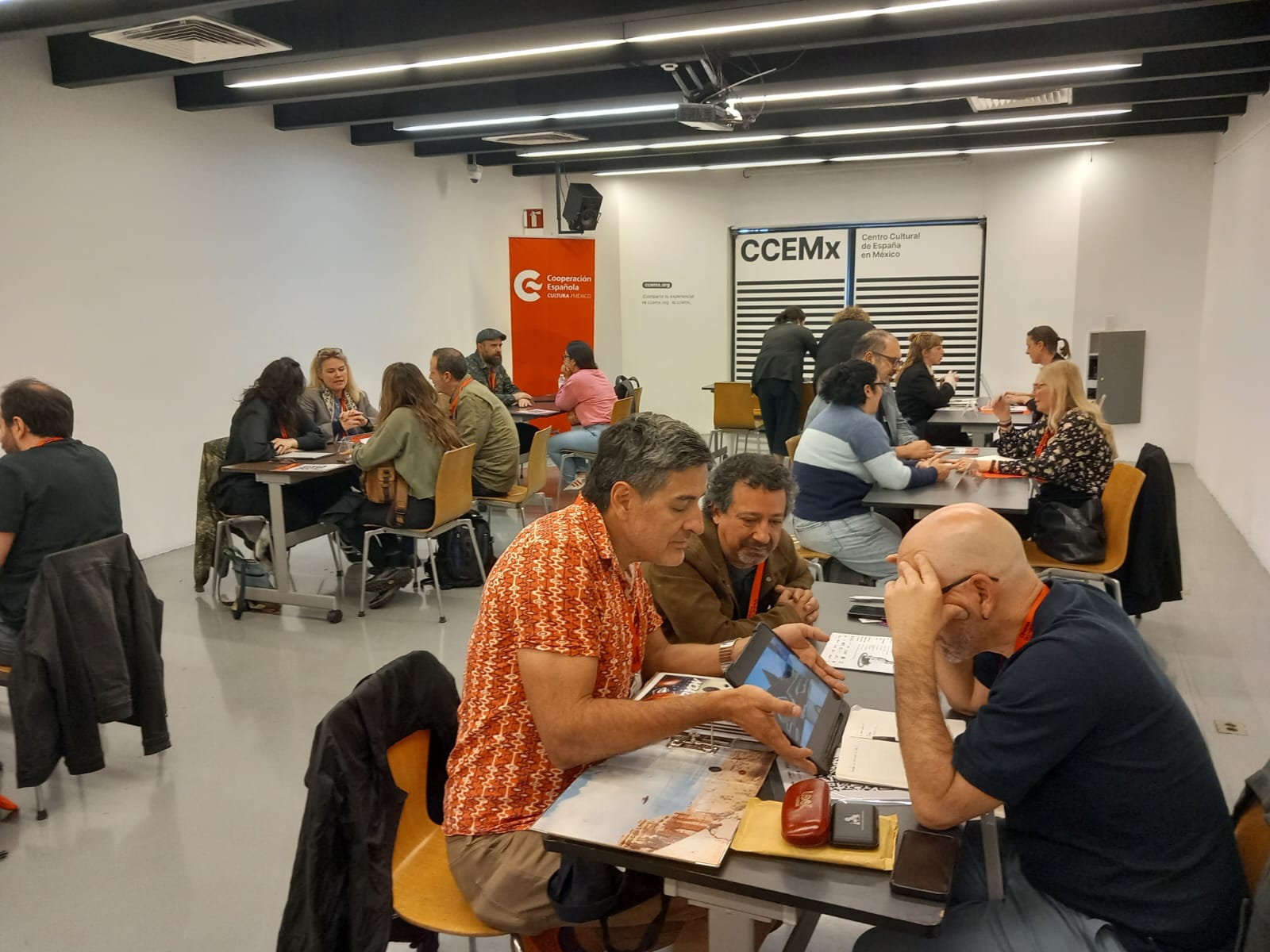 En primer plano dos personas comparten un proyecto audiovisual con otra en una mesa de reuniones. Al fondo se ven otros grupos de personas en el resto de mesas de reuniones.