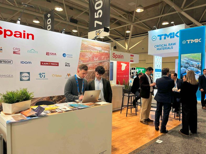 Dos personas revisan información en un portátil en el stand de una de las empresas participantes del Pabellón España de PDAC. Al fondo se ve a varios visitantes del Pabellón.