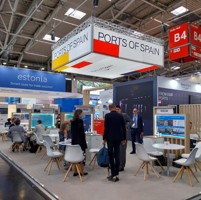 Varias personas se congregan en torno a las mesas de reuniones del sector de Ports of Spain, dentro del Pabellón España de Transport Logistic.