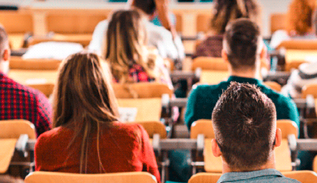 Varios alumnos de espaldas sentados en los asientos de un aula universitaria atienden a una clase.