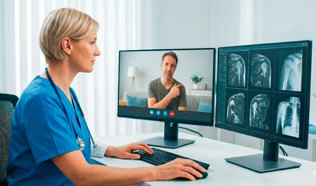 Una médico revisa radiografías en un monitor mientras atiende a un paciente por videollamada en otro monitor.