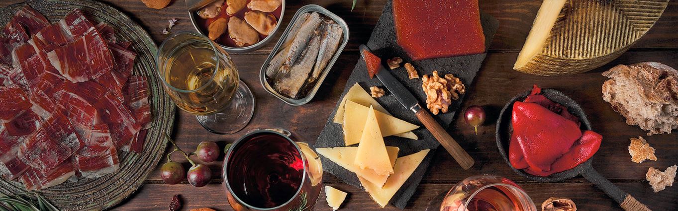 Un cotillón que incluye cuñas de queso, una bandeja de jamón ibérico, copas de vino blanco y tinto y latas de conservas  