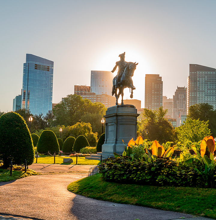 Estatua ecuestre en un parque urbano al atardecer, con jardines y rascacielos modernos al fondo.