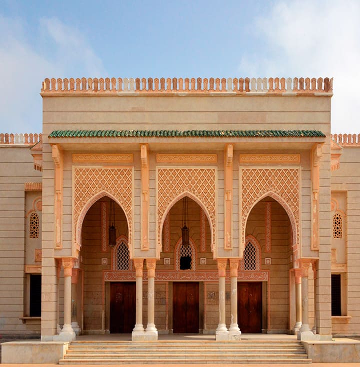 Fachada simétrica de un edificio de estilo islámico con tres arcos ornamentados, columnas delgadas, puertas de madera y escalinata frontal bajo un cielo azul.