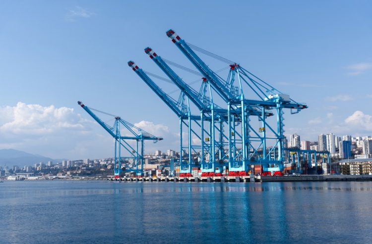 A large industrial container port featuring towering blue gantry cranes, stacked multicolored shipping containers and modern port infrastructure. Calm water in the foreground reflects the cranes, while a dense urban cityscape rises in the background. The scene is captured in Rijeka, Croatia, highlighting global trade, logistics, maritime transport and industrial infrastructure. Ideal for commercial use in topics such as shipping, import export, supply chain, freight operations, international business and economic development.