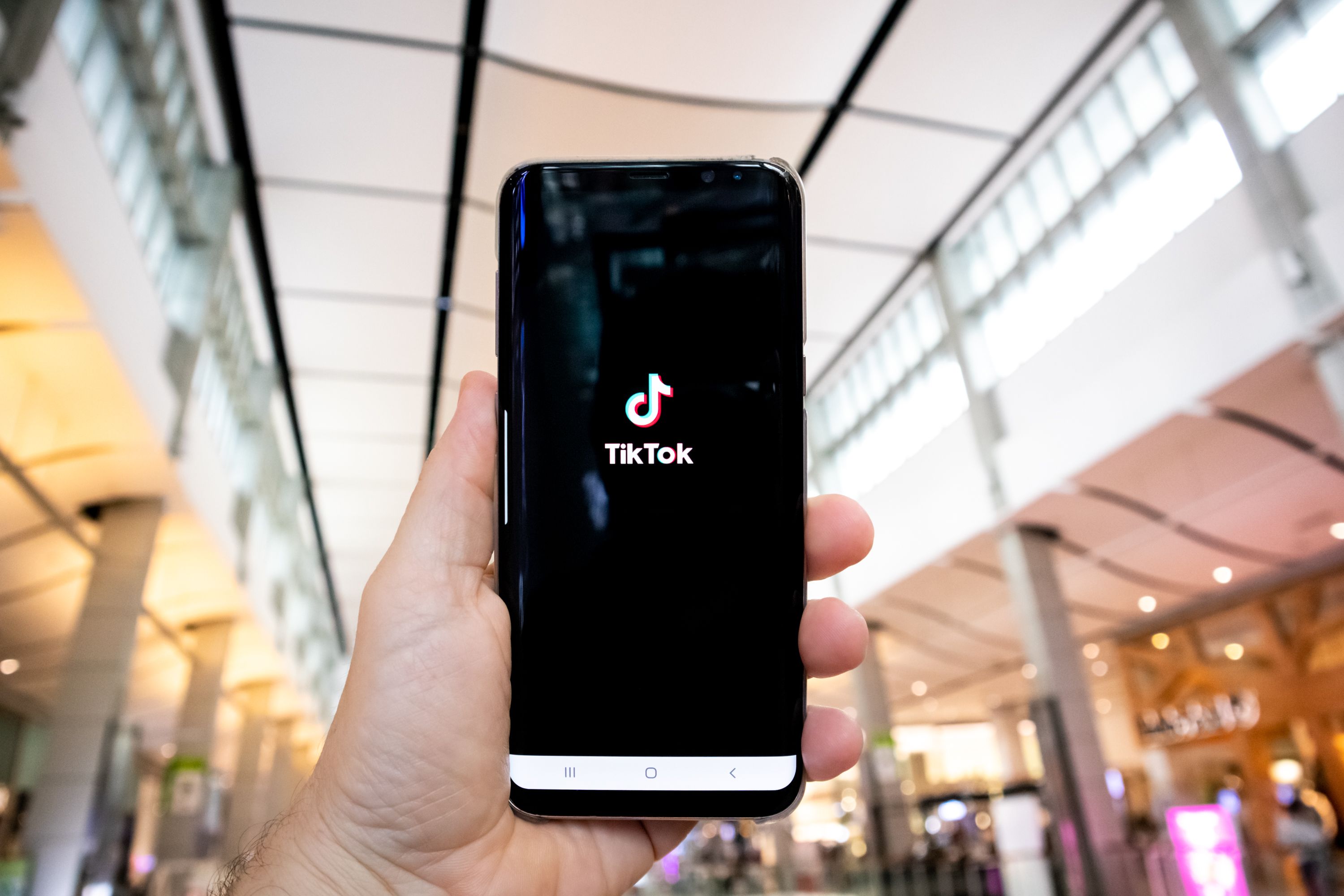 Imagen ilustrativa para la noticia "Singapur impulsa nuevos empleos gracias a TikTok Shop". Muestra una mano sosteniendo un móvil con la pantalla de carga de TikTok y un centro comercial de fondo.