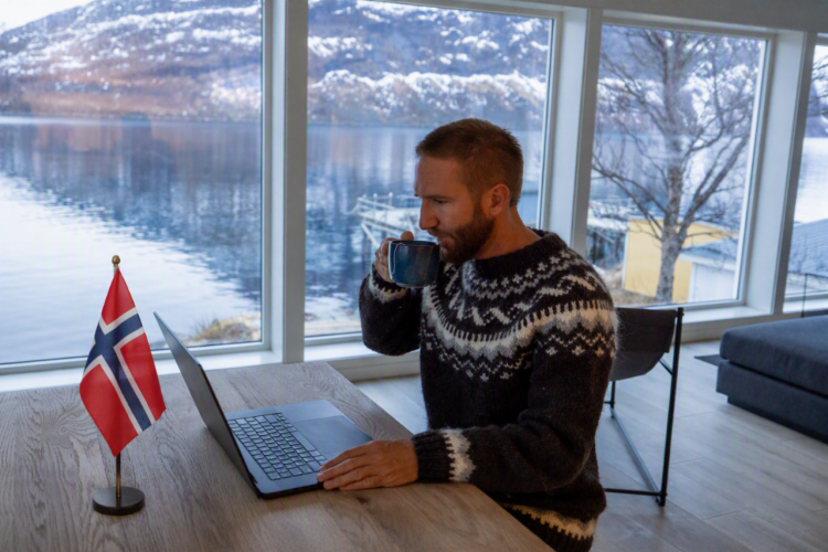 Imagen ilustrativa para la noticia "Claves de Noruega para el eCommerce nórdico y la compra transfronteriza". Muestra un hombre bebiendo de una taza mientras utiliza un ordenador portátil, con un lago de fondo y una bandera de Noruega en la mesa.