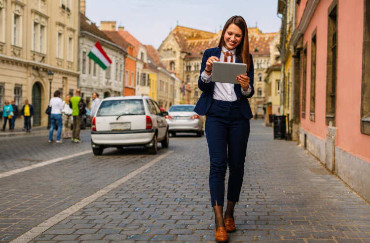 Imagen ilustrativa para el artículo "Hungría consolida el comercio electrónico con un récord de facturación en 2025 y un mercado en transformación". Muestra una mujer caminando por una calle en Hungría con una tablet en la mano.