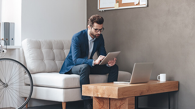 Multitasking. Handsome young man wearing glasses and working with touchpad while sitting on the couch in office